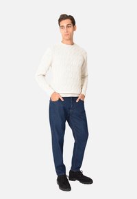 Maglione bianco lavorato a maglia con un motivo esagonale, pantaloni in denim blu e scarpe nere stringate, in piedi di fronte a uno sfondo neutro.