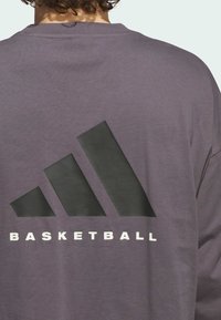 Felpa viola con una grande grafica nera di tre strisce diagonali e il testo "BASKETBALL" in bianco, posizionato sul retro.