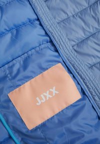 Interno della giacca imbottita blu con texture trapuntata. Presenta un'etichetta in tessuto pesco con "JJXX" in testo bianco. Bordo con zip di un blu contrastante.
