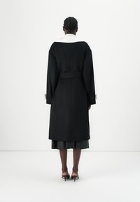 Manteau long noir avec un col blanc, taille ceinturée et manches larges. Texture lisse avec une coupe droite, dispose de poignets à bouton.