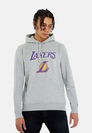 Giovane uomo con capelli intrecciati che indossa una felpa con cappuccio grigio chiaro con il logo dei Lakers viola e giallo, in piedi su sfondo bianco.