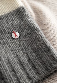 Grijze gebreide stof met ribbelstructuur, omrand door crème en lichtroze secties. Heeft een ronde grijze label met een rode silhouet van een hond.