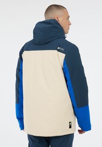 Farbig blockierte Jacke mit einem beigen Körper, navyblauen und leuchtend blauen Ärmeln, einer Kapuze und Logo-Details auf der Rückseite. Sanfte Textur.