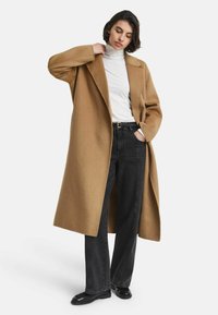 Vrouw die de kraag van een lange camelcoat aanpast, in een witte coltrui, zwarte hoge taille jeans, zwarte riem en zwarte schoenen voor een witte achtergrond.