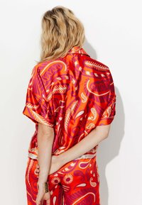 Chemise en satin rouge à manches courtes, avec un motif abstrait vibrant en orange et violet. Texture lisse et coupe décontractée.