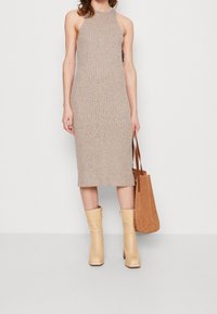Beige klänning i stickad struktur, ärmlös design, med liv hög slits, i kombination med bruna läderstövlar och en brun tote-väska.
