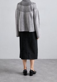 Harmaa ribbineulottu neule, jossa on korkea kaulus, pidettynä mustan polvenmittaisen hameen päällä. Mustat paksupohjaiset korolliset kengät täydentävät asun, nähtynä takaa.