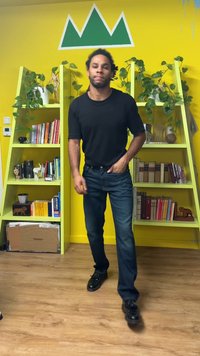 Musta lyhythihainen paita, siniset farkut ja mustat kengät. Taustalla vihreät kasvihyllyt ja keltainen seinä, joissa koriste-esineitä.