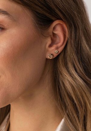 Nahaufnahme eines Frauenohrs mit zwei goldenen Blumen-Steckohrringen, einem größeren unten und einem kleineren oben, teilweise sichtbar sind hellbraune Haare.