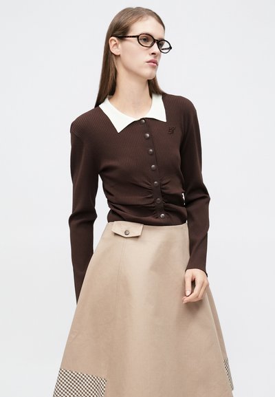 Καφέ ζακέτα ριμπ με λευκή γιακά και κουμπιά στο εμπρός μέρος· συνδυασμένη με μπεζ φούστα που έχει λεπτομέρεια με καρό σχέδιο.