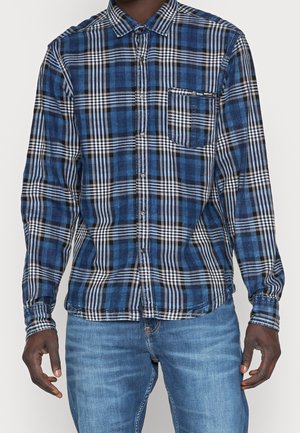 Hombre que lleva una camisa de manga larga a cuadros azul, negra y blanca con bolsillo en el pecho y vaqueros azules.