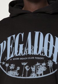 Černá mikina s kapucí s velkým modrým textem "PEGADOR" a grafikou palem s nápisem "MIAMI BEACH CLUB RESORT" pod tím.