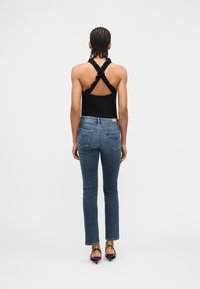 Άτομο με πλεκτά μαλλιά φοράει μαύρο τοπ με βολάν και χιαστί λουράκι, μπλε τζιν παντελόνι και μαύρες γόβες με ανοιχτή πλάτη, στέκεται γυρισμένο με την πλάτη προς μια λευκή επιφάνεια.