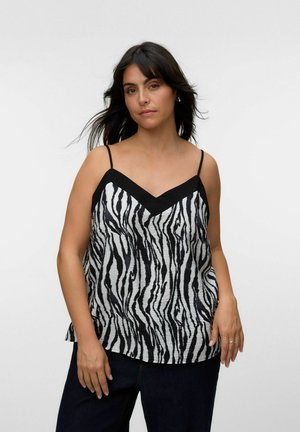 Femme aux longs cheveux foncés porte un débardeur noir et blanc à motif zèbre avec de fines bretelles et un pantalon noir, debout devant un fond uni.