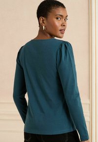 Une chemise à manches longues turquoise avec un col rond et des détails de manches bouffantes. Le tissu semble doux avec une texture lisse.