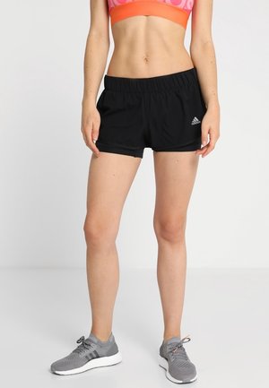 Vrouw draagt zwarte Adidas sportbroek, roze-oranje sportbeha en grijze hardloopschoenen, staand tegen een effen achtergrond.