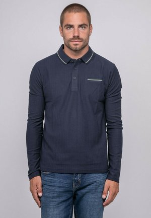 Homme aux cheveux courts et barbe, portant un polo bleu marine à manches longues et un jean bleu, debout devant un fond uni gris clair.