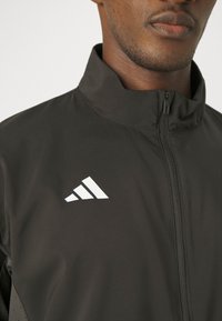 Czarna sportowa kurtka z wysokim kołnierzem, z białym logo Adidas na klatce piersiowej i teksturowanym materiałem oraz zapięciem na zamek.