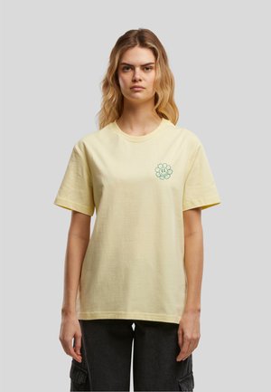 Femme aux cheveux blonds ondulés portant un t-shirt jaune pâle avec un petit motif de fleur souriante verte sur le côté gauche de la poitrine et un pantalon noir.