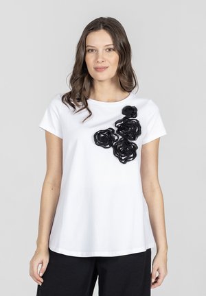 Kobieta o falistych brązowych włosach, ubrana w białą koszulkę z krótkim rękawem ozdobioną na piersi czterema czarnymi kwiatami z tkaniny, w połączeniu z czarnymi spodniami.