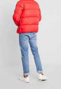 Rød pufferjakke med quiltet design, denimblå jeans og hvide sneakers. Pufferen har en høj krave og lynlås i siden.