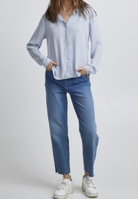 Camicia azzurro chiaro a maniche lunghe con chiusura a bottoni e colletto, abbinata a jeans di denim a vita alta e sneakers bianche.
