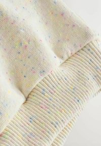 Tela de color amarillo claro con motas multicolores. La textura es de punto con un detalle de puño acanalado. Material suave y acogedor.