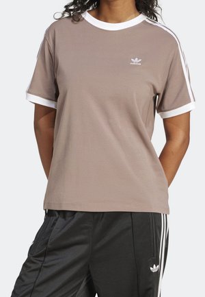 Person iført en lysebrun Adidas t-shirt med hvid kant og logo, parret med sorte Adidas træningsbukser med hvide striber og logo.