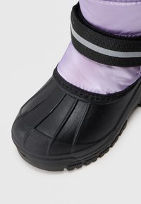 Semelle en caoutchouc noir avec des rainures, tige en nylon violet, maintenue par une sangle noire avec une bande grise réfléchissante.