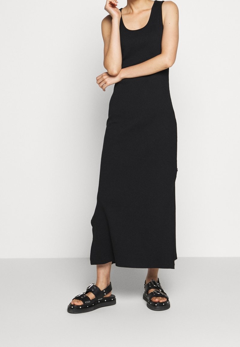 Femme portant une robe maxi noire sans manches et des sandales noires cloutées, debout les bras croisés devant un fond uni.