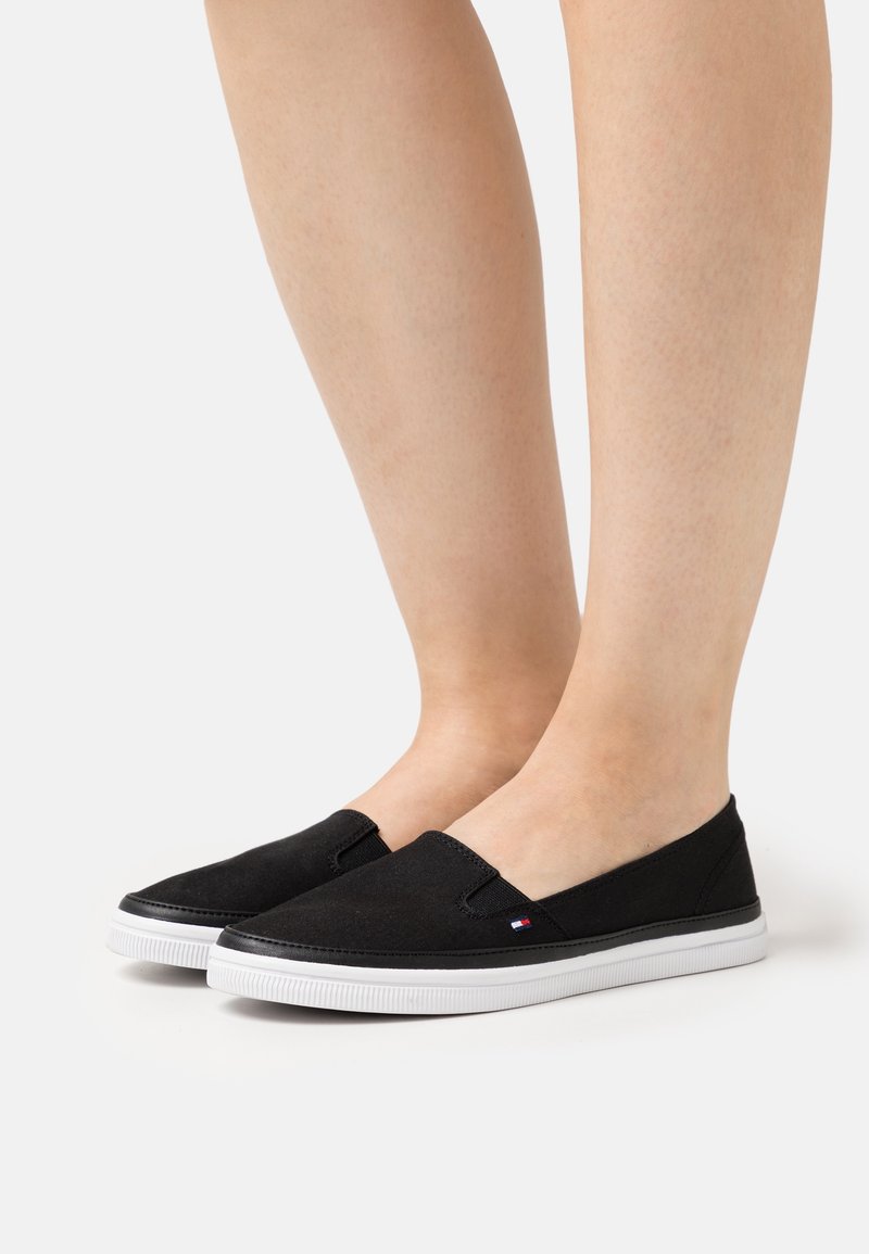 Pieds portant des chaussures en toile noires à enfiler avec des semelles blanches, petit logo sur le côté, posés sur une surface blanche.
