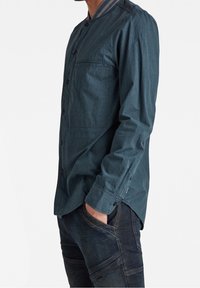 Camisa de manga larga verde oscuro con botones en la parte delantera, un bolsillo en el pecho y cuello en contraste. La tela parece suave con un acabado texturizado.