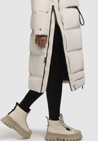Beige puffer coat with a zip closure, featuring a quilted design. Paired with black leggings and beige chunky-soled boots with laces.