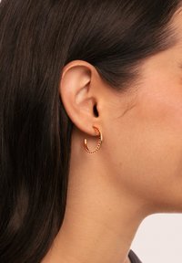 Boucle d'oreille en or ornée de pierres précieuses rouges le long du bord extérieur, mettant en valeur une finition lisse et polie. Portée dans un piercing à l'oreille.