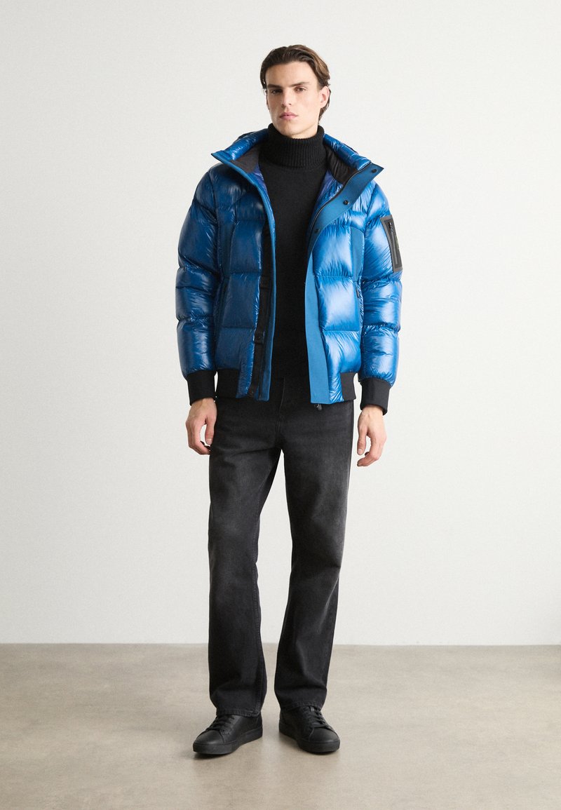 Doudoune bleue avec une finition brillante, dotée d'une capuche et d'une fermeture éclair à l'avant. Un col roulé noir, un pantalon foncé et des baskets noires complètent la tenue.