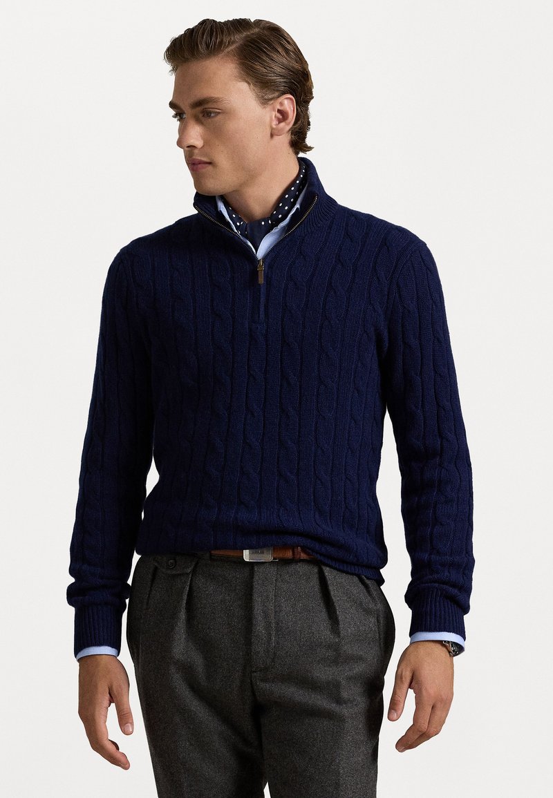 Μπλε σκούρο πουλόβερ με υφαντό πλεκτό σχέδιο και γιακά με φερμουάρ στη μέση, φορεμένο πάνω από ανοιχτό γαλάζιο πουκάμισο με κουμπιά, σε συνδυασμό με γκρι παντελόνι και ζώνη.