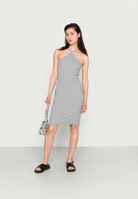 Vestido cinza, justo, com decote halter e design de alças cruzadas. Combinado com sandálias pretas e uma bolsa branca com um padrão abstrato preto.