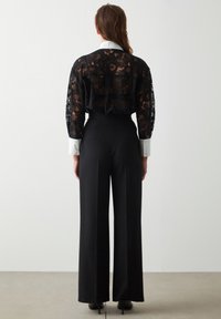 Kobieta ubrana w czarne szerokie spodnie oraz czarną koronkową bluzkę z długim rękawem i białymi mankietami, stojąca twarzą do białej ściany.