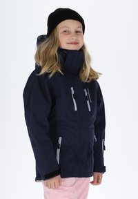 Granatowa wodoodporna kurtka z wysokim kołnierzem, licznymi kieszeniami na zamek, szarymi akcentami i luźnym krojem, noszona na różowych spodniach.