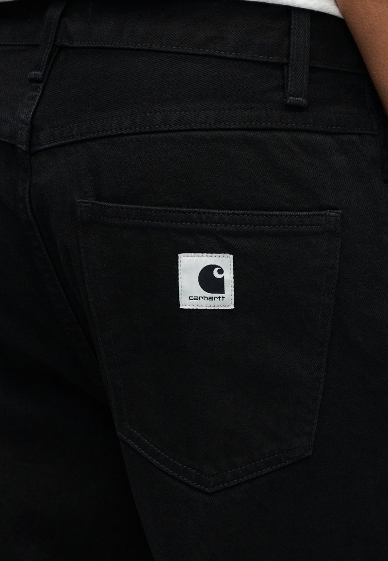 Zwarte spijkerbroek met een achterzak voorzien van een wit label met een logo. De stof heeft een gladde textuur met zichtbare stiksels.