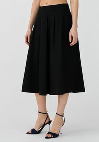 Gonna nera plissettata con vita aderente, lunghezza al ginocchio, realizzata in un tessuto liscio. Abbinata a sandali con tacco blu navy.