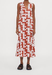 Maxiklänning med ett geometriskt mönster i vitt och rött, en djup V-halsringning och ärmlös design, kombinerad med svarta sandaler.
