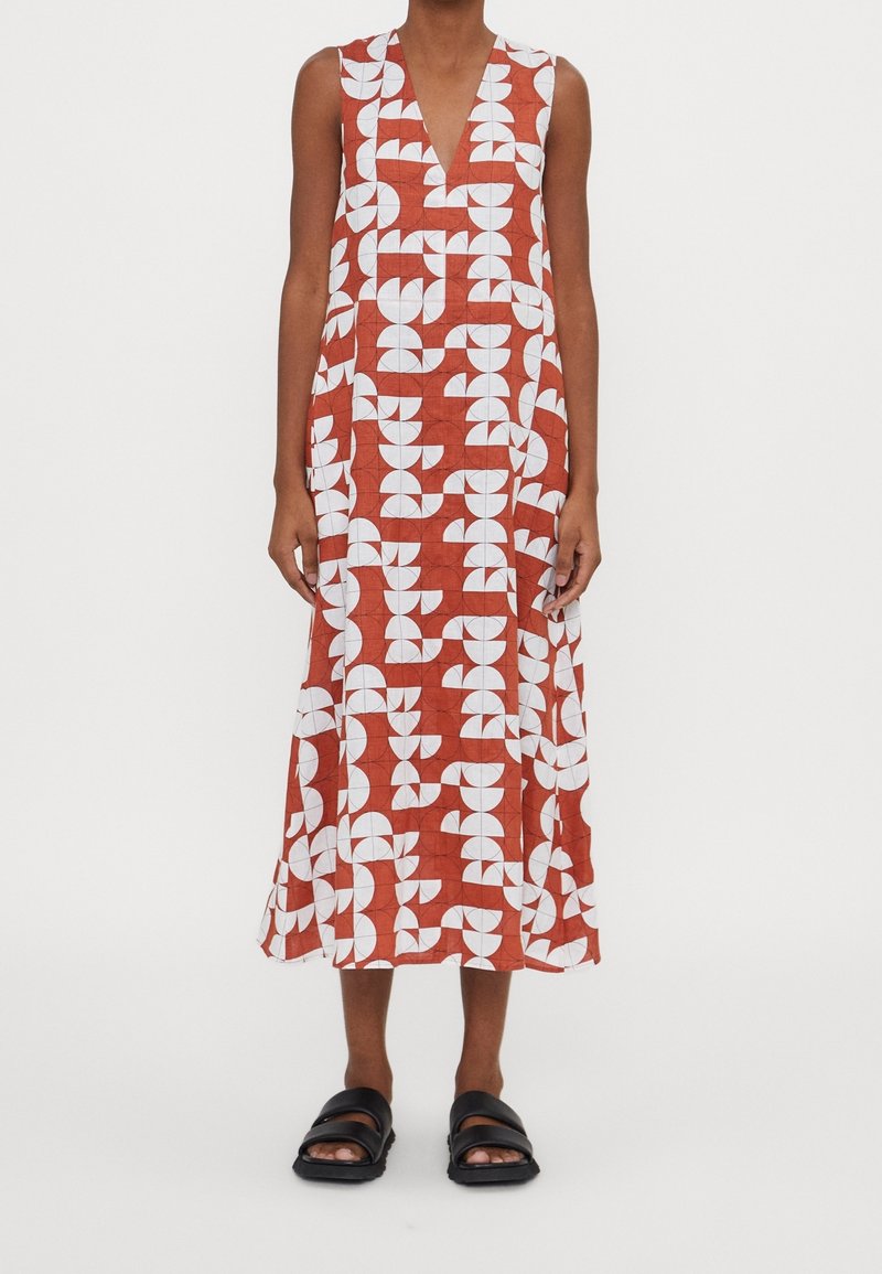 Maxiklänning med ett geometriskt mönster i vitt och rött, en djup V-halsringning och ärmlös design, kombinerad med svarta sandaler.