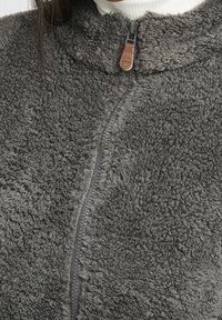 Harmaa fleece-takki, jossa on vetoketju edessä, pehmeä ja teksturoitu pinta sekä ruskea nahkakoristevetoketjun vetimessä.