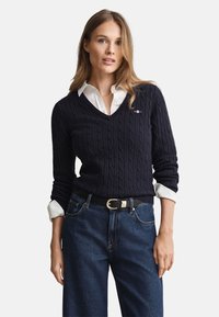 Marineblauer Strickpullover mit V-Ausschnitt, getragen über einem weißen Hemd mit Kragen. Kombiniert mit dunkler Jeans und einem schwarzen Gürtel mit silberner Schnalle.