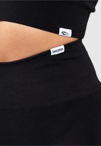 Zwarte geribde sporttop en legging met logo-labels. Het materiaal lijkt rekbaar en aansluitend, met een elegante uitstraling en strakke lijnen.