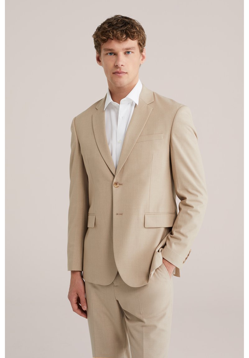 Giovane uomo con capelli ricci, indossa un completo beige con una camicia bianca, in piedi con una mano in tasca contro uno sfondo chiaro e semplice.