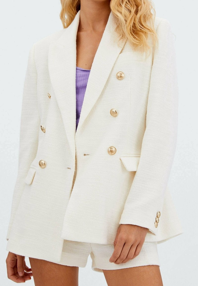 Femme portant un blazer blanc texturé à double boutonnage et un short assorti avec des boutons dorés, sur un haut violet.