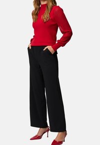 Czerwona sweter z prążkowanymi bufkami po bokach w zestawie z czarnymi szerokimi spodniami. Modelka ma na sobie czerwone czółenka na szpilkach; ręce w kieszeniach.