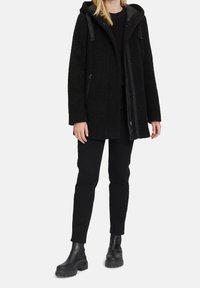 Abrigo negro texturizado con capucha, cierre frontal y bolsillos laterales. Lleva puesto un suéter negro y combina con pantalones negros y botas gruesas negras.